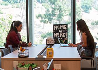 Two students studying together at a table. A Home is Here sign in the background.