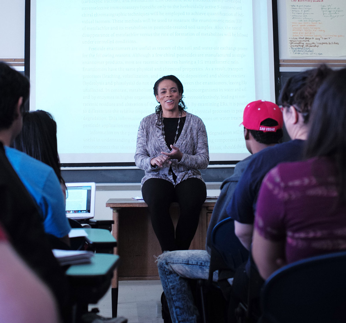 A faculty member sitting on a desk speaking to their class.