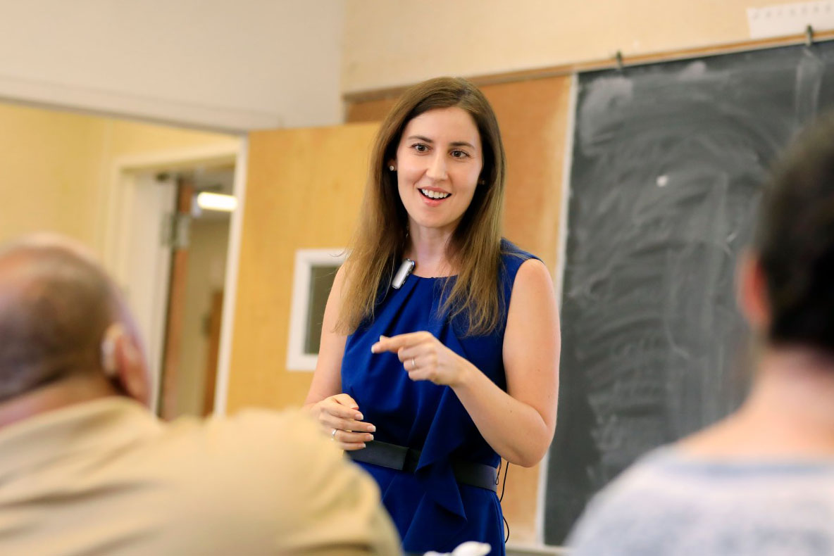 A person stands in a classroom. They are standing and appear to be speaking with two seated people.