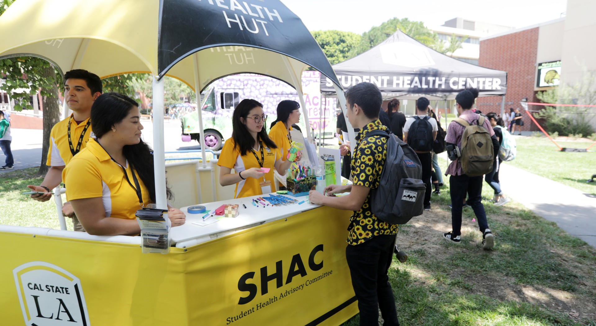 Student in a sunflower print shirt stands at a domed hut that is operated by three students wearing matching SHAC polos.