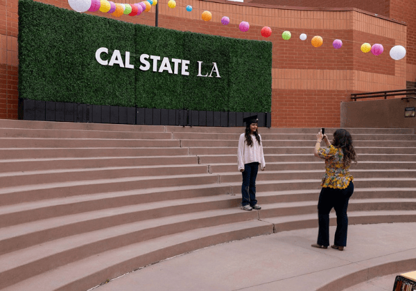 Youth wearing a graduation cap posing in front of a hedge with a Cal State LA sign as onlooker takes their photo