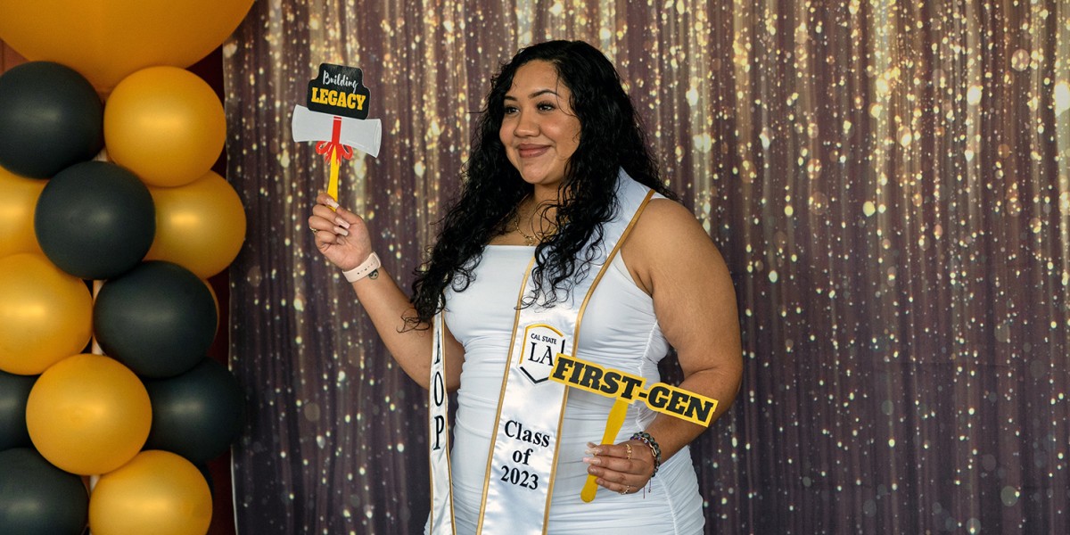 A student poses with "First Gen" sign in front of a glittery curtain and balloons.