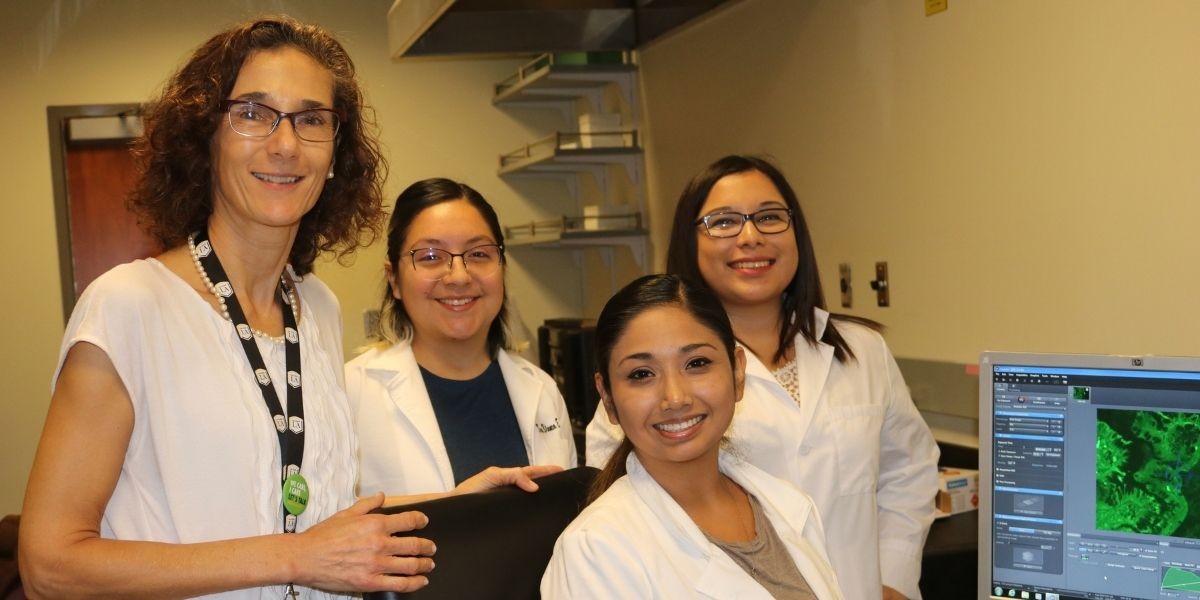 Three students in white coat and professor in a lab. Computer monitor displays image of organism