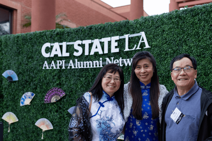 Three people standing in front of a green hedge decorated with fans from the Asian diaspora and a sign that says CAL STATE LA