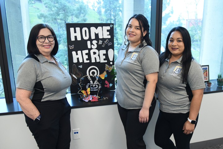 Three people smiling, standing around a sign that reads "Home is Here!"