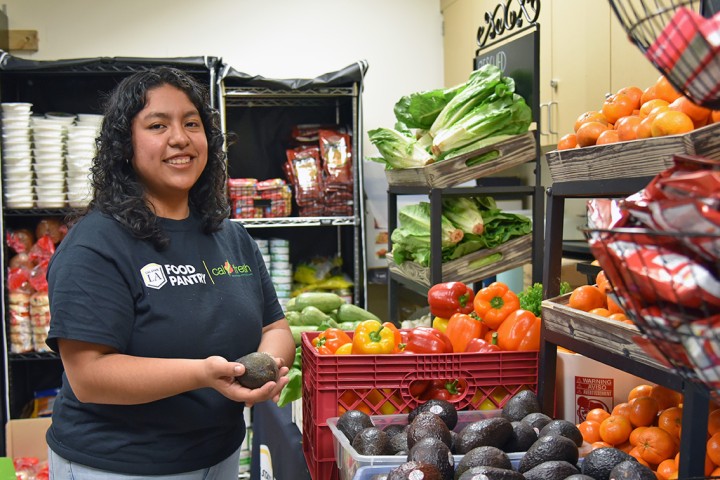 A person wearing a Cal State LA Food Pantry t-shirt smiling while standing in front of bins of fresh produce.