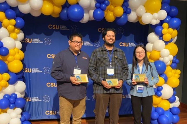Three students (2 male, 1 female) standing with their awards on a stage that is decorated with balloons