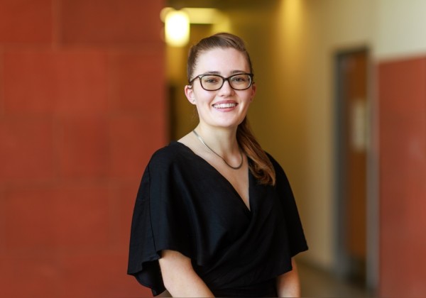 Researcher smiles while standing in a campus hallway, wearing a black top and glasses at Cal State LA.