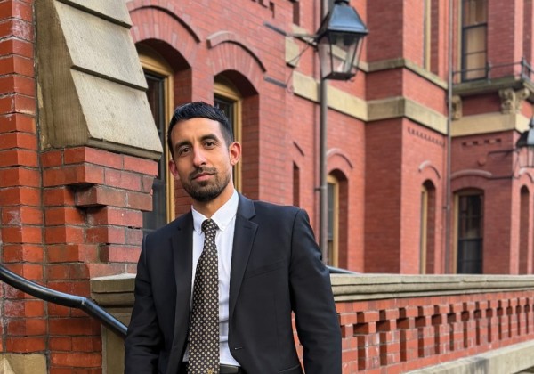 Individual in a dark suit stands outside a historic brick building, posing for a portrait. 