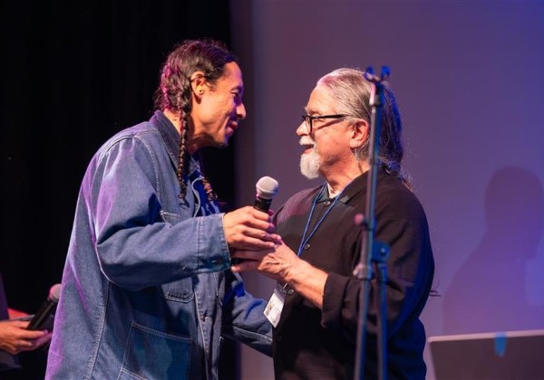 Two individuals greet and share a handshake on stage during the LAS 65 Years Celebration event at Cal State LA, under stage lighting with microphones visible.