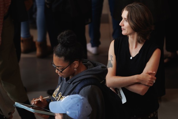 A diverse group of attendees gather, some taking notes and others engaged in conversation, in a large indoor event space.