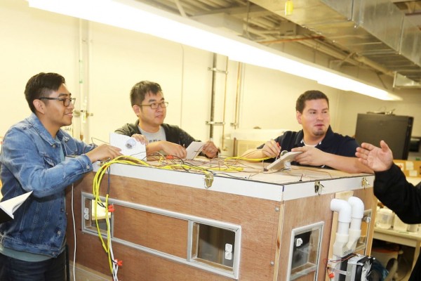 Image of a group of engineering students surrounding a large wooden box device while their professor addresses them. 