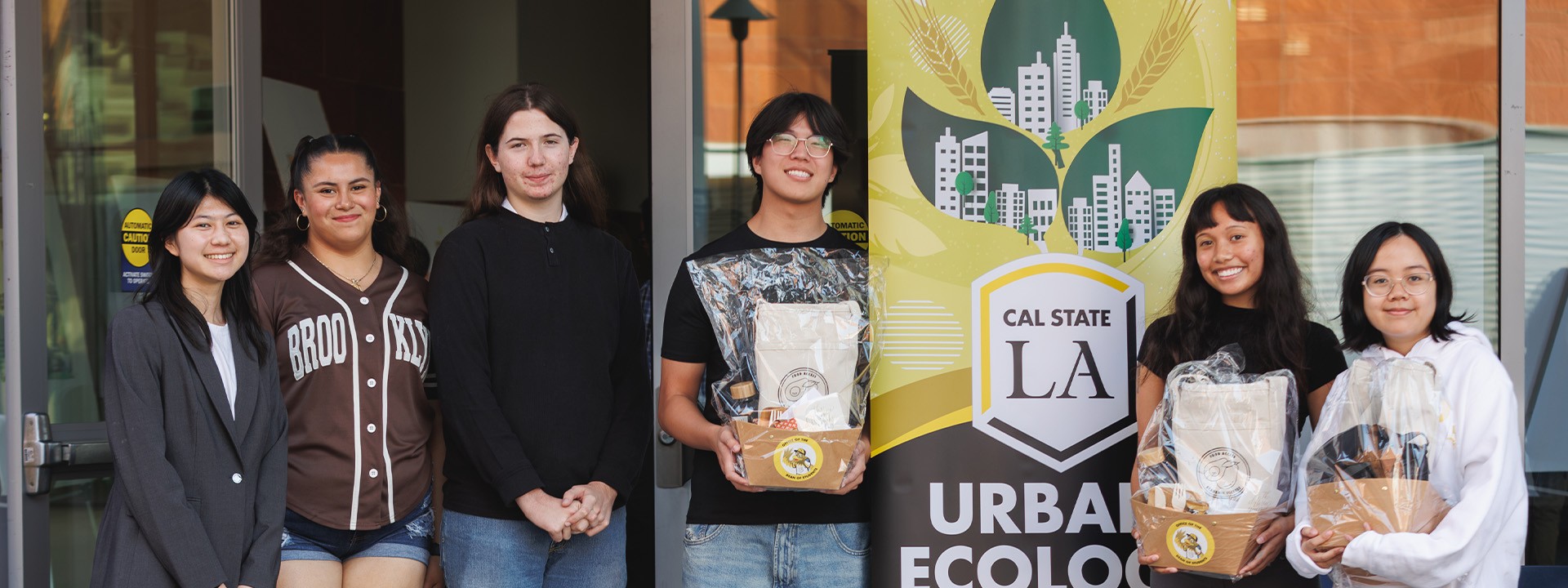 A group of students stands beside a Cal State LA Urban Ecology banner, holding gift bags and smiling at the camera.