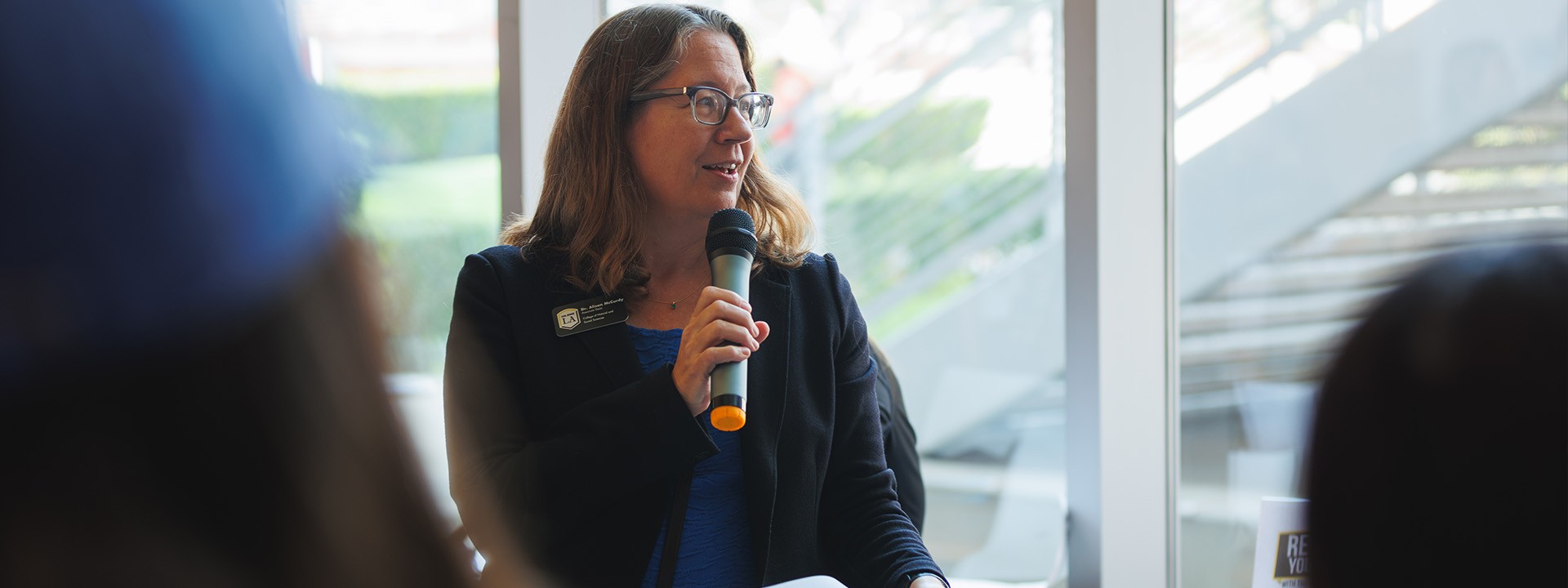 A woman with glasses, wearing a name tag, speaks into a microphone, likely addressing the audience.