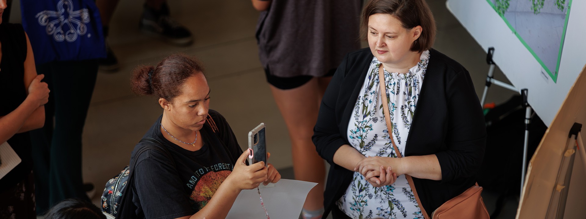 Two women observe event materials, with one taking a photo and the other looking on with interest.