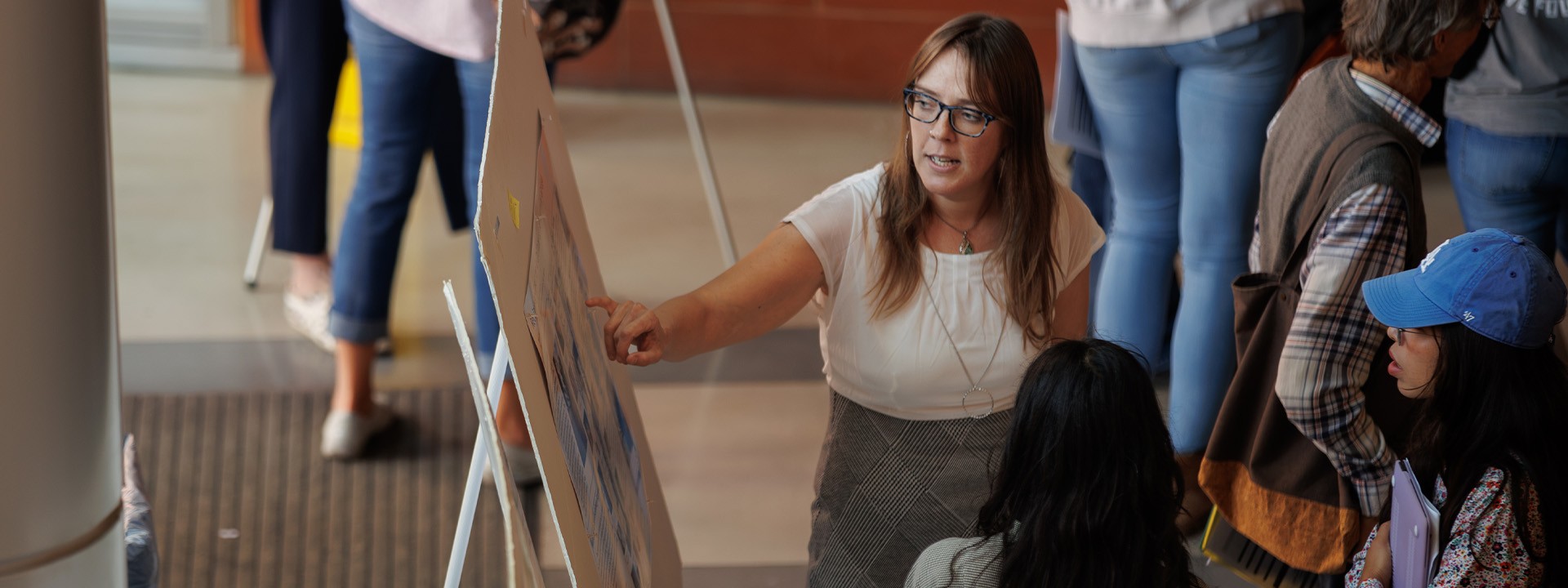 A woman points to information on a large poster board while talking to a small group of attendees.