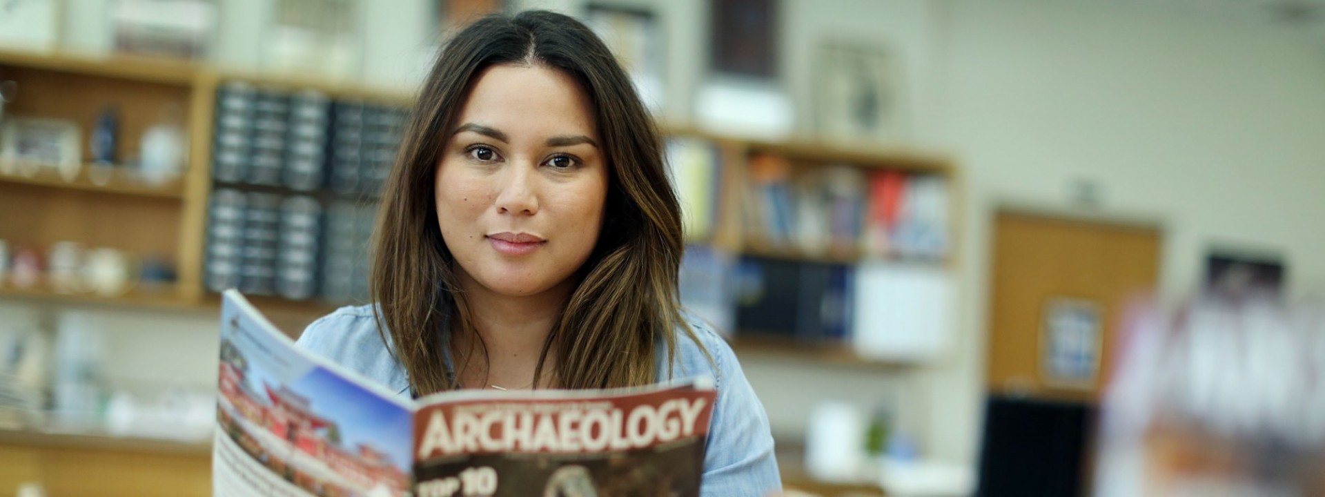 Image of a female student looking directly into the camera as she holds a copy of Archaeology Magazine in a classroom. 