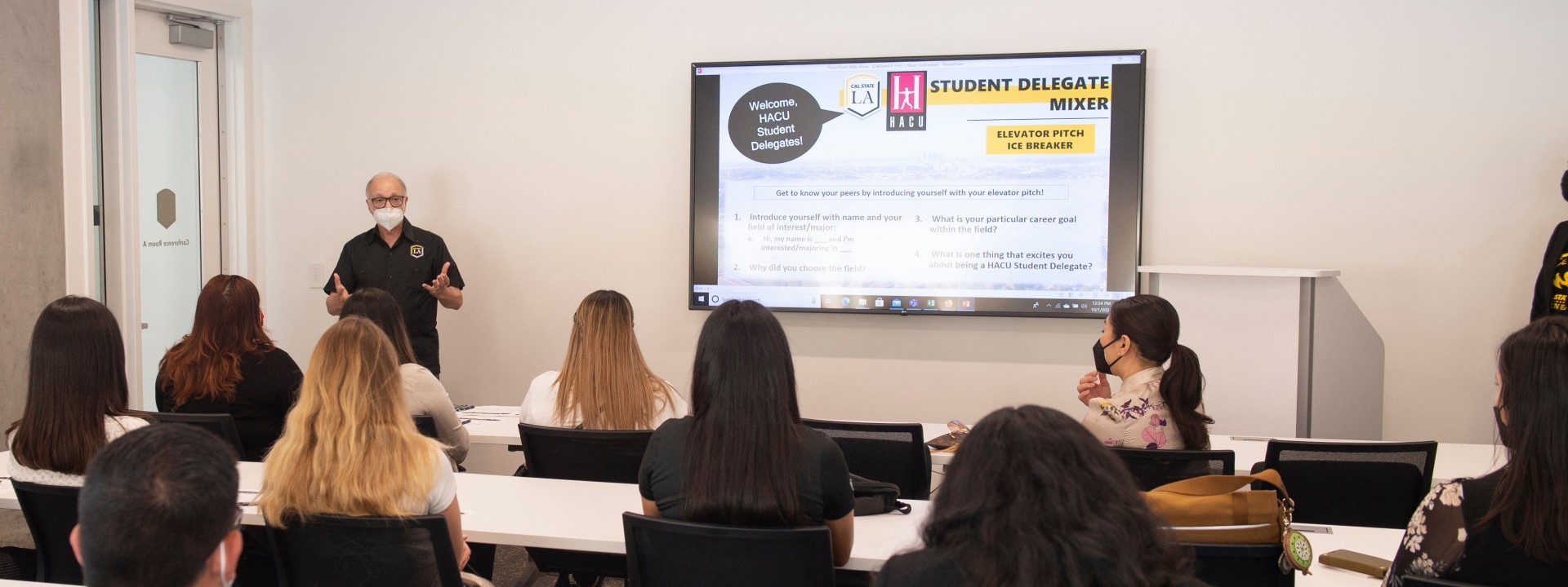 Person wearing a mask speaking in front of a class, standing next to a smart screen.